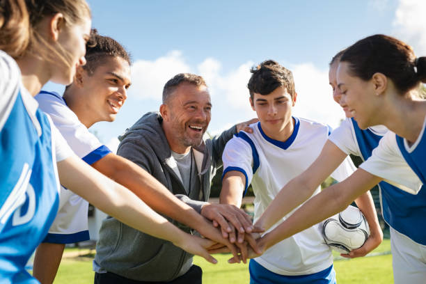 Happy coach and high school soccer team with their hands stacked in football court. Young football players stacking hands together. Mature man coach encourages his students to do their best during the sport match.